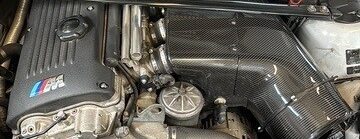 Ein detaillierter Blick auf einen Motorraum eines Autos mit sichtbaren Motorkomponenten und einer Abdeckung aus Kohlefaser.