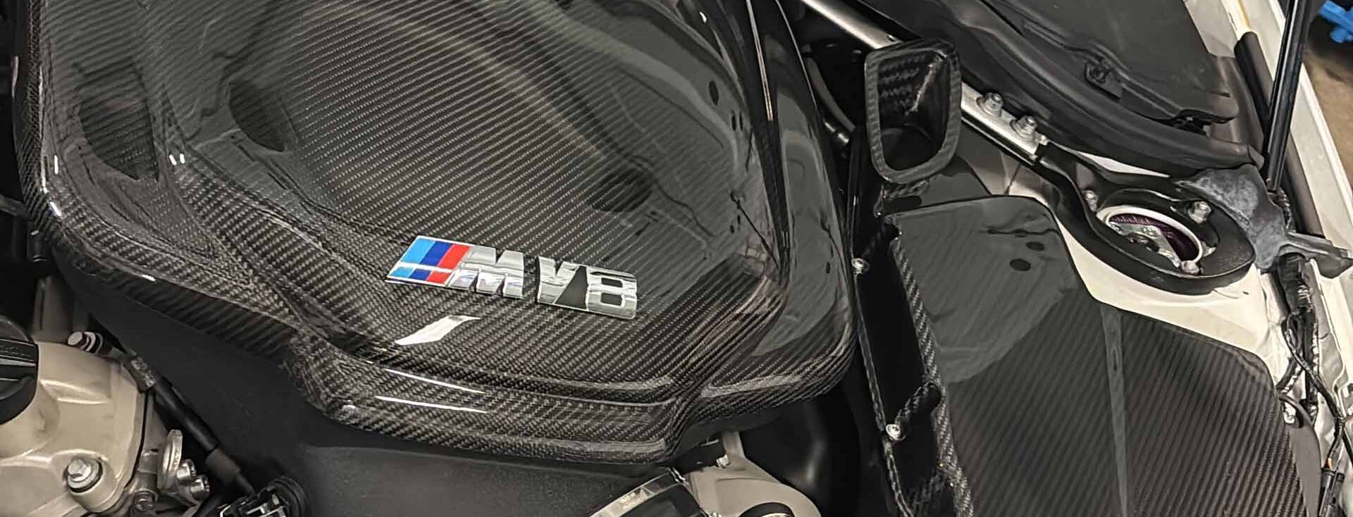 Motorraum eines Wagens mit schwarzer Kohlefaserabdeckung und M-Logo.